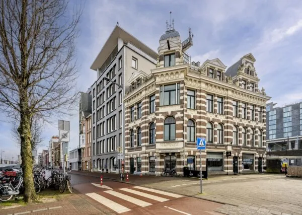 Straatbeeld van de De Ruijterkade in Amsterdam met een historisch hoekpand en moderne bebouwing op de achtergrond.